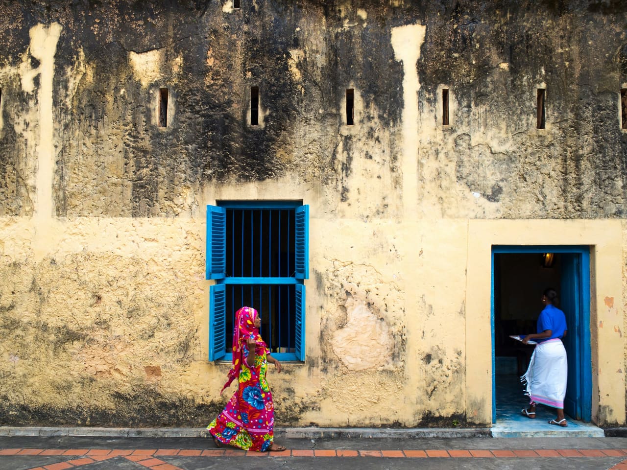 A woman in a brightly patterned dress walks past a weathered coral-stone wall with blue shutters in Stone Town, Zanzibar